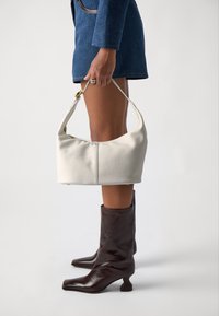 Sac à épaule en cuir blanc avec une finition texturée, affichant une silhouette courbée et des accents en métal doré, tenu par une personne vêtue d'une tenue bleue.