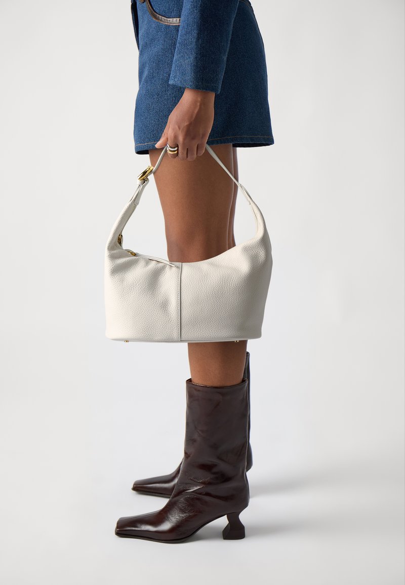 Sac à épaule en cuir blanc avec une finition texturée, affichant une silhouette courbée et des accents en métal doré, tenu par une personne vêtue d'une tenue bleue.