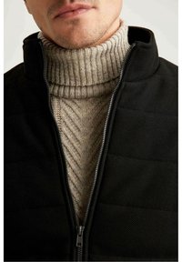 Svart vadderad jacka med hög krage, öppnad för att avslöja en beige ribbad polotröja under. Texturerade mönster synliga på tyget.