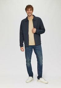 Marineblaue Zip-Jacke über einem beigen Shirt, kombiniert mit blauen Jeans und weißen Sneakers. Die Jacke hat einen flachen Kragen und gerippte Bündchen.