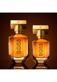 Deux flacons de parfum en verre ambré avec des bouchons rectangulaires dorés, étiquetés "BOSS" et "THE SCENT", présentés sur une surface réfléchissante.