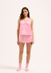 Ensemble de pyjama en satin rose comprenant un débardeur à fines bretelles et un col arrondi, assorti à un short et des chaussettes roses en peluche.