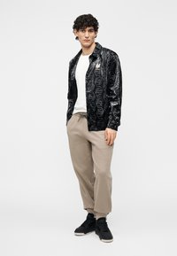 Čierna lesklá bunda s hexagonálnym vzorom, nosená cez bielu košeľu a béžové tepláky. Má detail s logom a čierne tenisky.