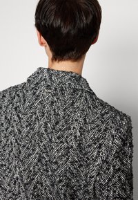 Person mit kurzem dunklem Haar, von hinten zu sehen, die eine schwarz-weiße strukturierte Fischgrätenjacke mit einem kleinen "BOSS"-Logo am Kragen trägt.