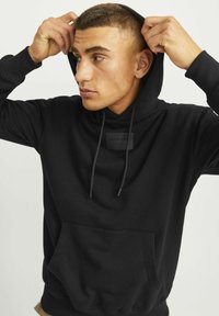 Sudadera negra con capucha, bolsillo frontal, cordones y una etiqueta de logo texturizado en el pecho, hecha de material suave.