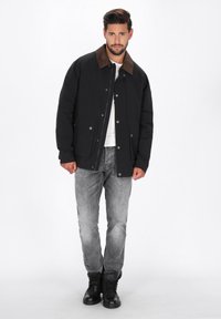 Sort jakke med brun krave, knaplukning og to forlommer, båret over en hvid skjorte og parret med grå jeans og sorte støvler.