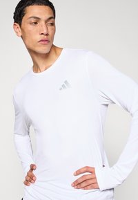 Biała sportowa koszulka z długim rękawem o żebrowanej fakturze i zaokrąglonym dekolcie, z odblaskowym logo Adidas na lewej piersi.