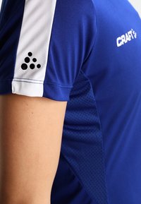 Blå atletisk skjorte med en hvid ærme, der har et sort prikmønster logo. Materialet er glat, med et struktureret mesh-panel for åndbarhed.