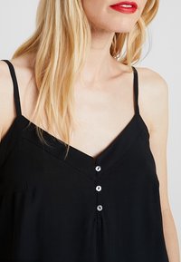 Blond kvinna som bär en svart ärmlös topp med tunna band och tre små knappar i mitten, röd läppstift synligt, ljus hudton.