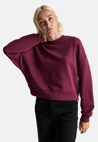 Sudadera cortada burdeos con cuello redondo, corte relajado y puños acanalados. Combinada con jeans de tiro alto negros. Textura suave.