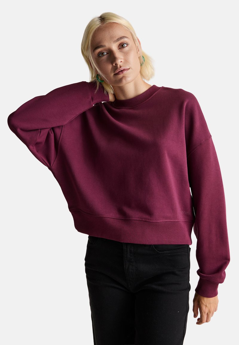 Bordowy, skrócony sweatshirt z okrągłym dekoltem, luźny krój i ściągacze przy rękawach. Noszony z czarnymi, wysokimi dżinsami. Gładka tekstura.