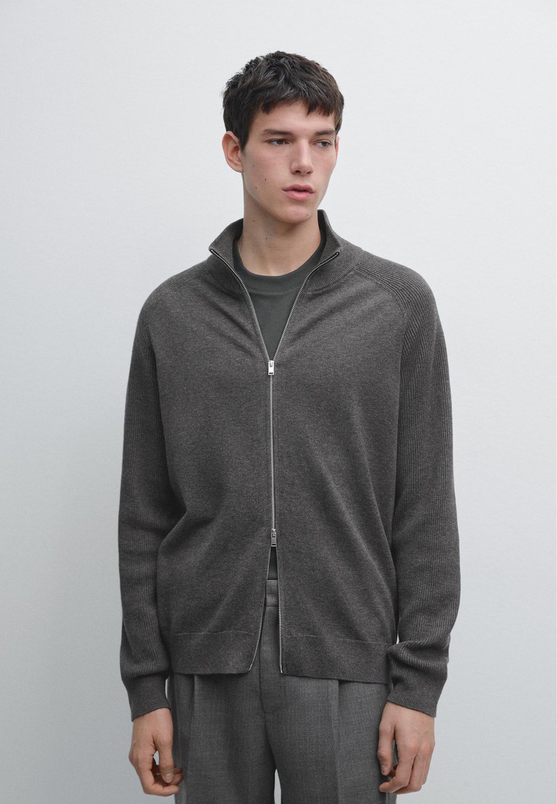 Jeune homme portant un pull à fermeture éclair intégrale gris anthracite sur une chemise sombre et un pantalon gris, debout devant un fond uni gris clair.