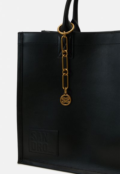 Sac fourre-tout en cuir noir avec un accent en chaîne dorée et un charme logo, texture lisse et nom de la marque en relief à l'avant.