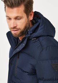 Marineblaue Steppjacke mit Kapuze, ausgestattet mit einer strukturierten Oberfläche, kontrastierenden Akzenten und einem kleinen Logo auf der linken Schulter.