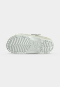 Σόλα λευκού clog Crocs που δείχνει υφή πέλματος με μοτίβο και σημάδια μεγέθους σε απλό ανοιχτό γκρι φόντο.