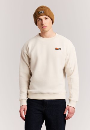 Jonge man die een beige fleece trui en een bruine gebreide muts draagt, staand tegen een effen lichte achtergrond.