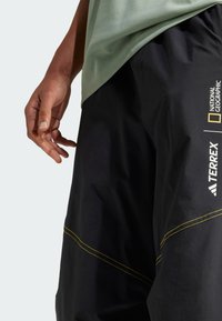 Melni āra šorti ar dzelteniem izšuvumiem un National Geographic un Terrex logotipiem sānos, kopā ar zaļu kreklu.