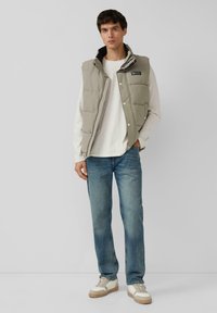 Hellgraue Steppweste mit hohem Kragen; mit einem schwarzen Etikett, silbernen Druckknöpfen und gestepptem Material ausgestattet. Kombiniert mit hellblauen Jeans und cremefarbenen Sneakers.