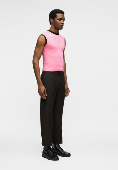 Άνδρας που στέκεται φορώντας ένα ροζ αμάνικο τοπ με μαύρη επένδυση, μαύρο ραμμένο παντελόνι και μαύρα μποτάκια με κορδόνια σε ουδέτερο φόντο.