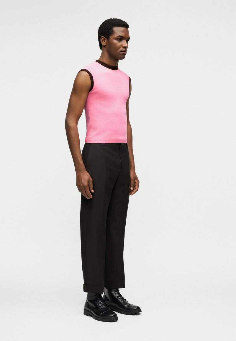 Man standing wearing a sleeveless pink top with black trim, black tailored trousers, and black lace-up boots against a plain background.