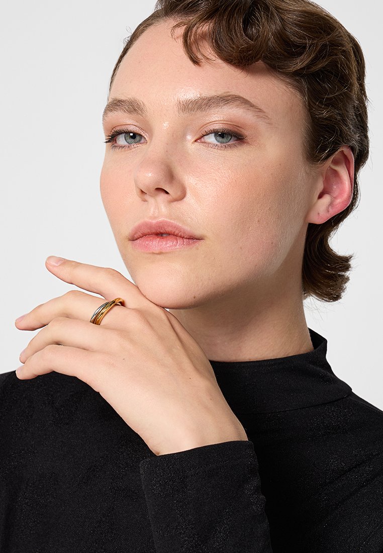 Bague en or et en argent au doigt du modèle. Le modèle a les cheveux courts et porte un top noir à manches longues avec un col montant.