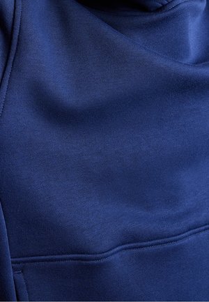 Sinisen collegepaidan kankaan lähikuva, jossa näkyvät ompeleet ja taskun yksityiskohdat.