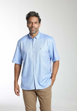 Homme aux cheveux gris portant une chemise à manches courtes bleu clair boutonnée et un pantalon beige, debout avec une main dans la poche devant un fond uni.