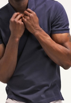 Mand iført en marineblå poloshirt, justerer kraven med begge hænder, hvilket viser muskuløse arme og en tatovering på højre arm.