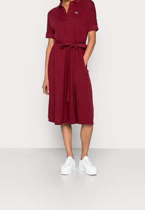 Vrouw draagt een bordeaux polo-jurk tot op de knie met een strikceintuur en witte platformsneakers, staand met één hand in de zak.