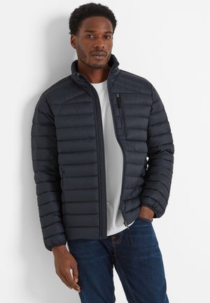 Man wearing a dark blue quilted jacket over a white shirt and blue jeans, standing with one hand in pocket against a white background.