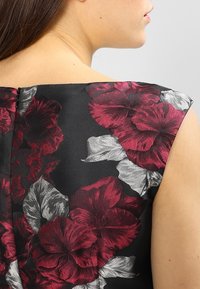 Květinové šaty s černým pozadím; obsahují velké květiny v burgundské a stříbrné barvě. Materiál má hladkou texturu; mezi detaily je také zip na zádech.