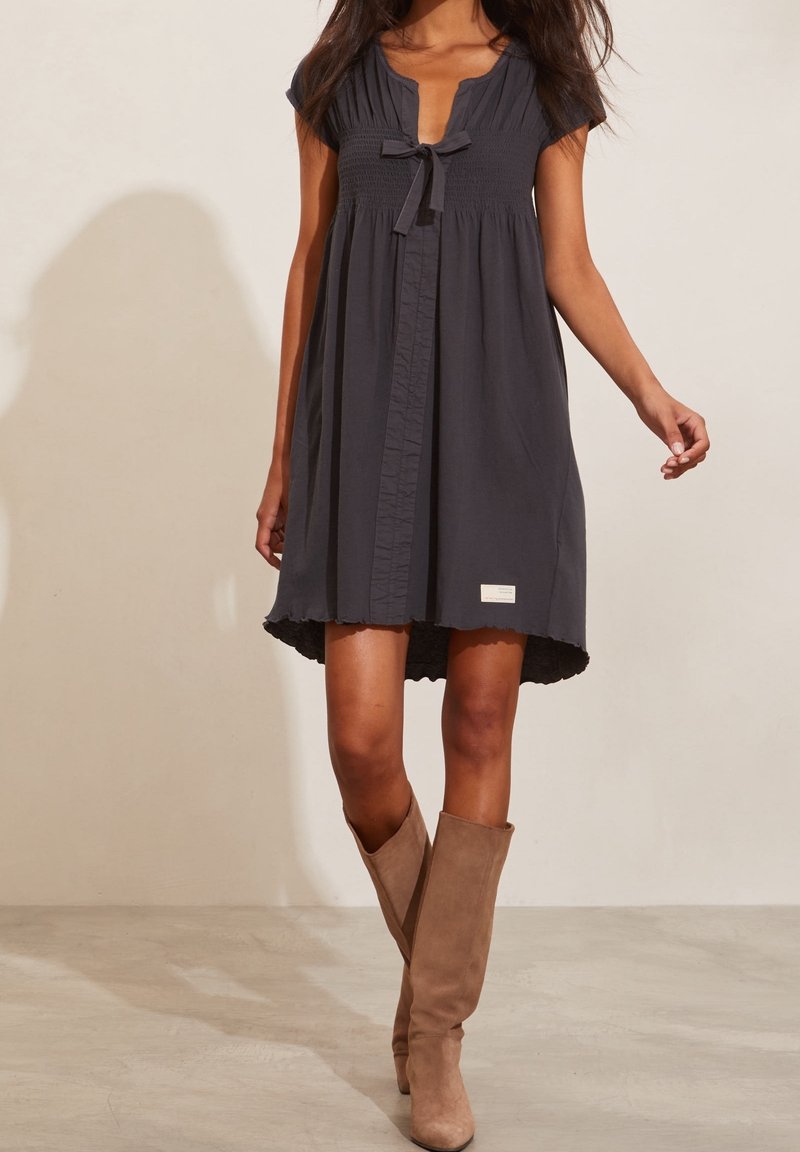 Tmavě šedé krátké šaty s krátkými rukávy, detaily na zavazování u výstřihu, knoflíková légou a vlnkovým lemem. Nosí se s krémovými kozačkami po kolena.