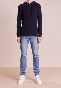 Námořnická modrá pletená mikina s kapucí a šňůrkou, v kombinaci s světle modrými džíny a bílými teniskami. Jednoduchý design, bez vzorů a ozdob.