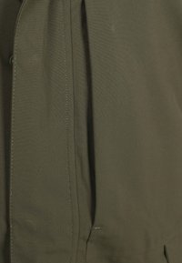 Olivengroene jas gemaakt van lichtgewicht stof, met zichtbare stiksels, een plooidetail en een discrete zijzak.