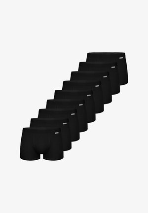 Trænet mand uden skjorte med lys hud og kort blondt hår iført sorte boxer shorts, stående mod en ensfarvet lys baggrund.