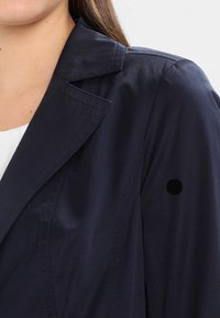 Mørkeblå trenchcoat med en struktureret krave, skuldersøm-detalje og en synlig sort knapaccent på højre ærme. Glat tekstur.