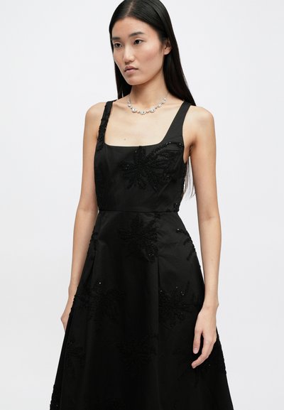 Μαύρο φόρεμα χωρίς μανίκια με τετράγωνη λαιμόκοψη και λουλουδάτη κέντημα με κοσμήματα. Απαλή ύφανση με φαρδιά φούστα. Διαυγής κολιέ για έμφαση.