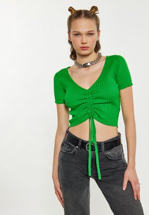 Jonge vrouw met twee space buns, gekleed in een groene gerimpelde cropped top, zwarte spijkerbroek, een dikke zilveren ketting en een zwarte riem.