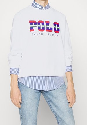 Bílá mikina s vícebarevnými pruhovanými nápisy "POLO" a "RALPH LAUREN", nošená přes modrobíle pruhovanou košili na knoflíky a světle modré džíny.