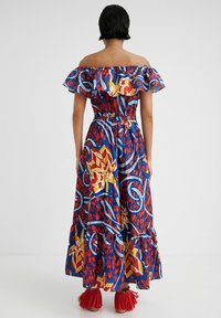 Maxi-jurk met off-shoulder ruffle-halslijn, vervaardigd uit een felblauwe, rode en gele stof met patroon. Flared zoom en bijpassende versierde sandalen.