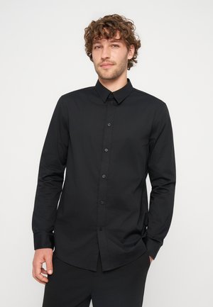 Homme aux cheveux bouclés portant une chemise noire ajustée à manches longues boutonnée, debout devant un fond clair uni.