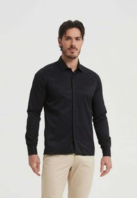 Chemise noire à manches longues avec un tissu lisse et un col pointu. Présente un ourlet droit et des boutons assortis. Portée avec un pantalon beige.