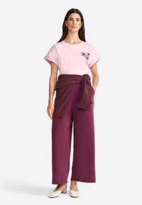 Camiseta de algodón color rosa claro con bordado floral, combinada con pantalones sueltos de color burdeos profundo y un suéter a juego drapeado en la cintura. Zapatos off-white.