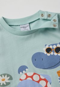 Vaaleansininen puuvillapaitapusero pyöreällä kaula-aukolla. Sisältää leikkisän hippo-grafiikan, joka käyttää aurinkolaseja, ja on korostettu värikkäillä kuvioilla ja kukkakoristeilla.