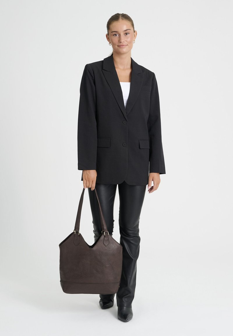 Femme portant un blazer noir et un pantalon, tenant un grand sac à main en cuir marron foncé avec deux anses d'épaule.
