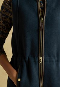 Sötétkék polár mellény arany cipzárral, barna díszítéssel és oldalsó zsebekkel. Textúrált anyag jellemzi, és állítható derékrésze van, így igényes fazont biztosít.