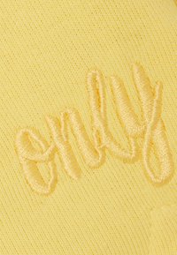 Tela amarilla tejida con una textura, bordado "only" en hilo contrastante. Textura sutil en toda la superficie, con una apariencia suave y transpirable.