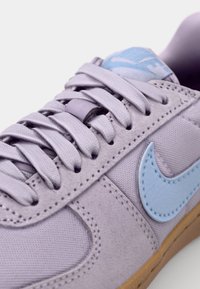 Baskets lavande clair avec une tige texturée, un swoosh Nike bleu et une semelle en caoutchouc marron. Comprend des lacets plats et une languette rembourrée.