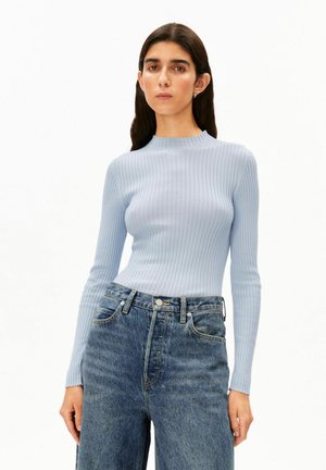 Femme portant un top côtelé à manches longues bleu clair rentré dans un jean bleu taille haute et coupe ample, debout devant un fond uni.