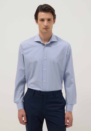 Man wearing light blue striped button-up dress shirt tucked into dark blue trousers, standing against plain light background.